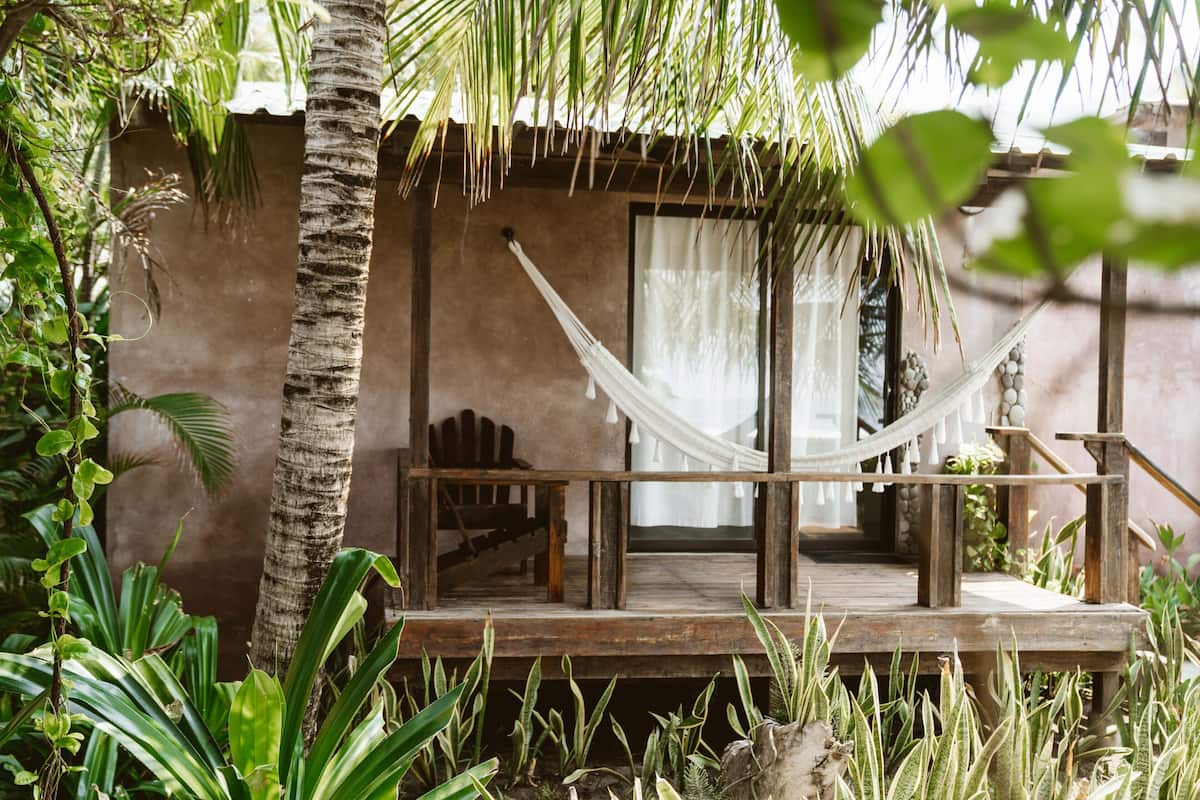 A wooden deck with a hammock, surrounded by tropical plants and a building with a large window.