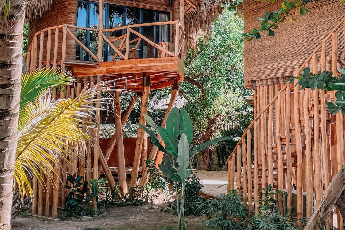 A thatched-roof wooden structure with a balcony, surrounded by lush greenery.