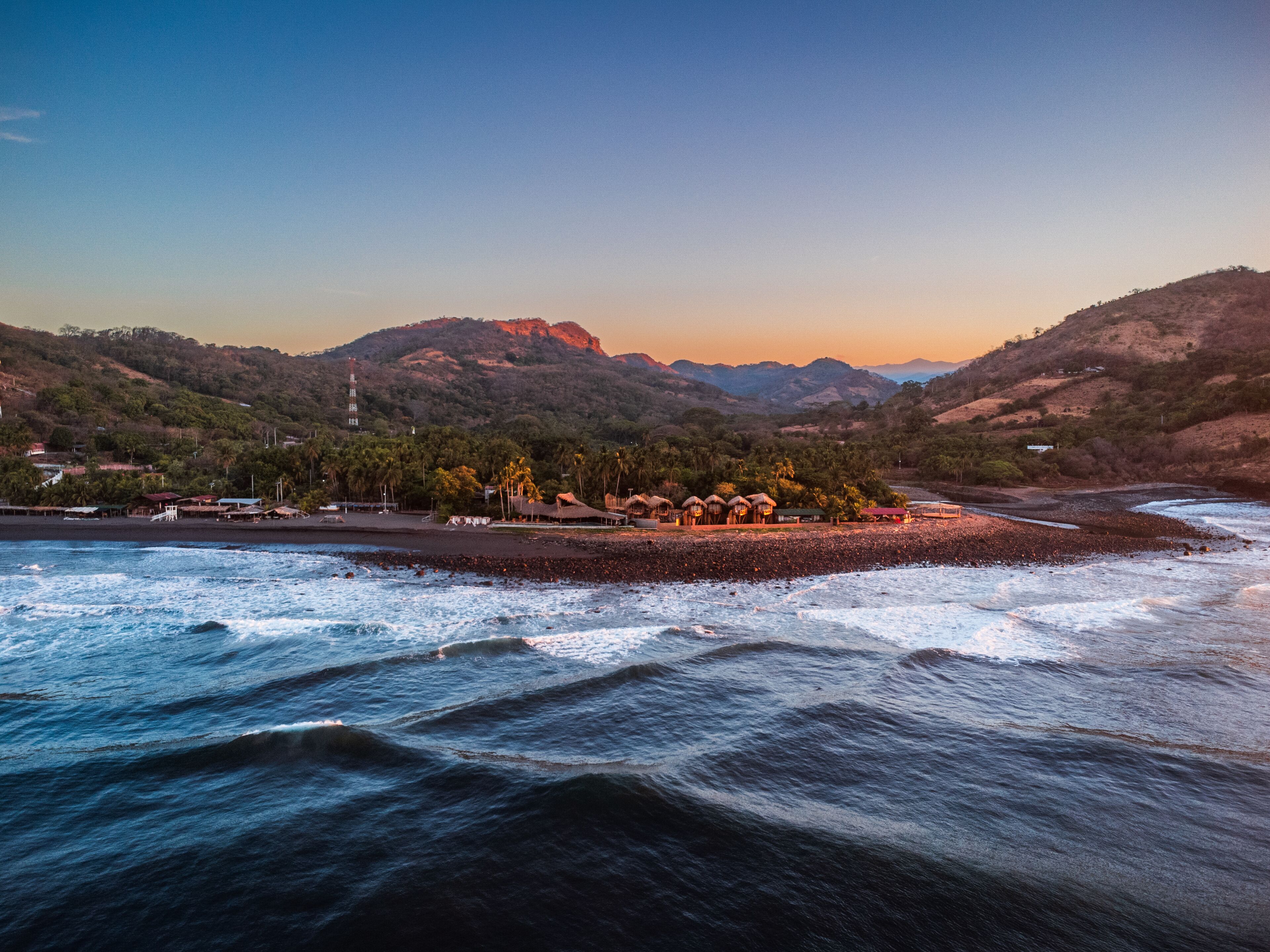 A coastal area with a beach, thatched-roof huts, and a mountain backdrop.