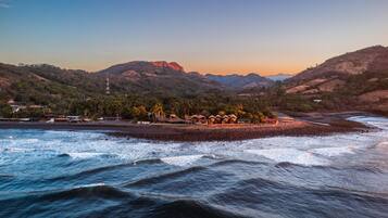 A coastal area with a beach, thatched-roof huts, and a mountain backdrop.