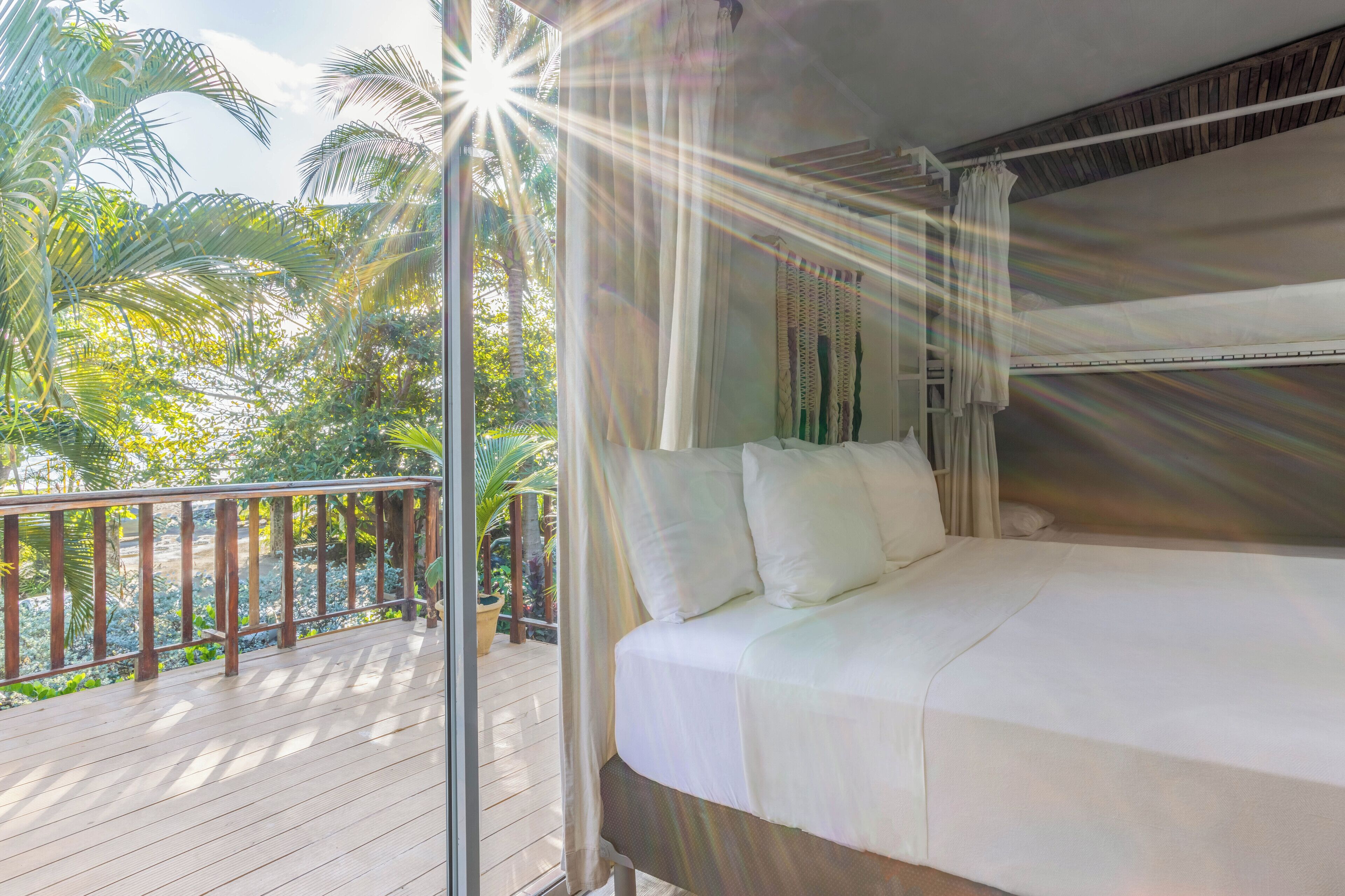 A balcony with a wooden railing, a bed with white bedding, and a view of tropical foliage.