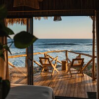A wooden deck with two chairs facing the ocean, a thatched roof, and a view of the sea.