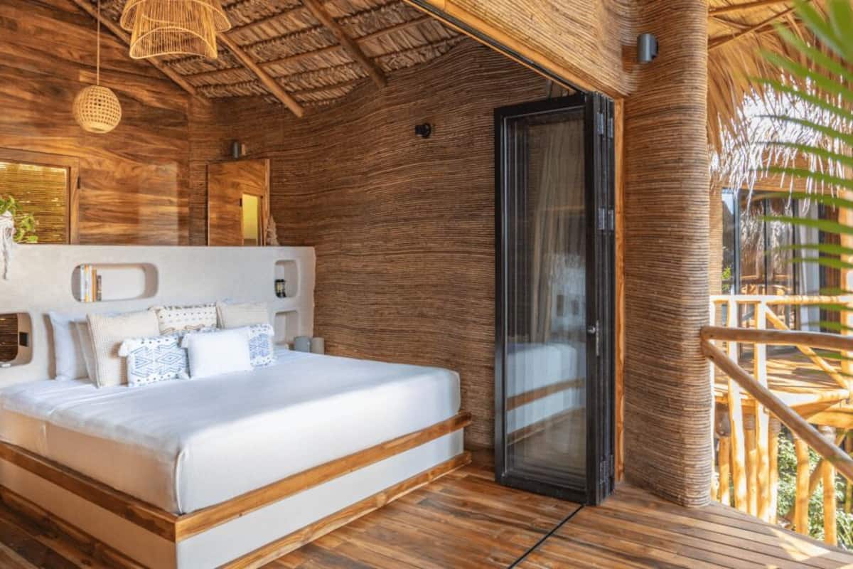 A wooden cabin-style bedroom with a large bed, a glass door leading to a balcony, and a woven ceiling.