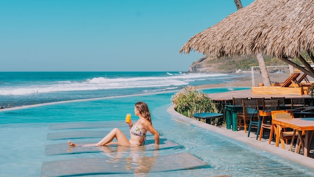 Una mujer descansando en una silla flotante en una piscina azul cristalina, con vistas al océano y palmeras.