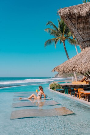A woman relaxing on a floating lounge chair in a clear blue pool with a view of the ocean and palm trees.