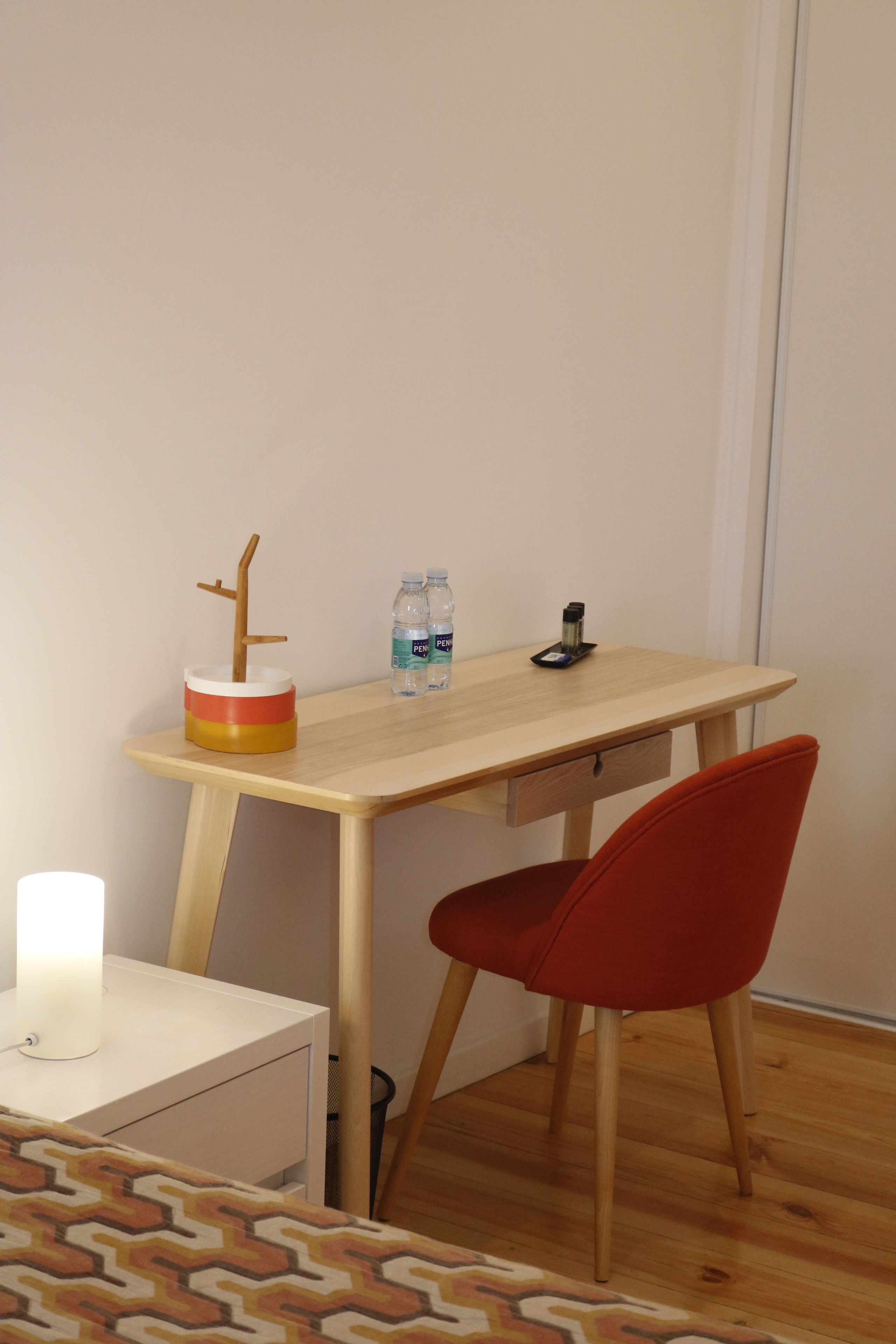 A small, neatly arranged room with a wooden desk, a red chair, a white bedside table, and a patterned rug.