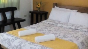 A neatly made bed with a floral bedspread, a yellow throw blanket, and two rolled towels. There is a small table with a vase of flowers beside the bed. - Zacona Eco-Resort and Biblical Garden (San Pablo)