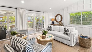 Una sala de estar moderna con un sofá, sillones, una mesa de centro y un comedor visible a través de puertas de vidrio.