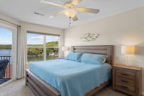 Ein Schlafzimmer mit Bett, Nachttischen, Deckenventilator, einem Fenster mit Blick auf ein Gewässer und einer Wanddekoration.