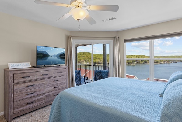 A bedroom with a bed, a television on a dresser, a balcony with a view of a lake, and a ceiling fan.