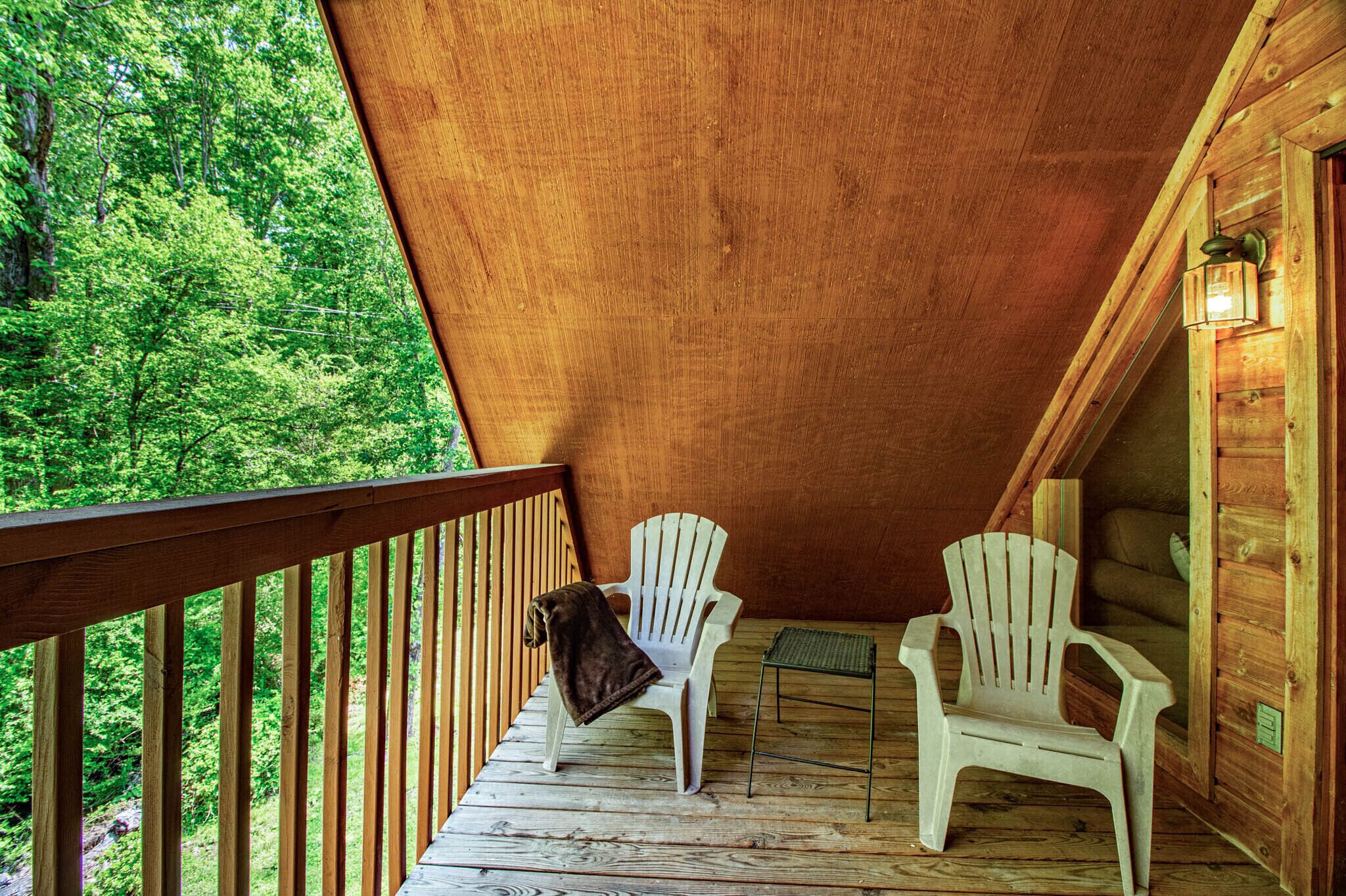 A wooden deck with two white chairs and a small table, overlooking a forested area.