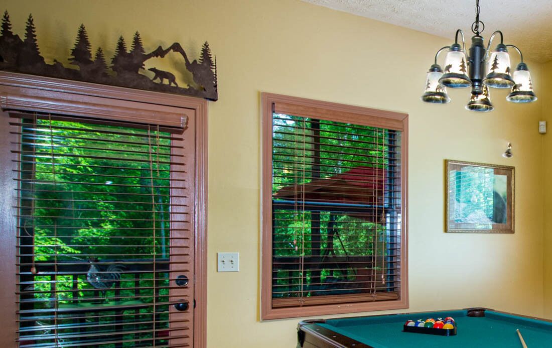 A pool table with a set of balls, a framed picture on the wall, and a window with blinds.