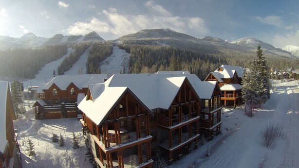A lodge with multiple wooden buildings, each with a snow-covered roof, set against a backdrop of snow-covered mountains and trees. -  Ski-In Ski-Out, Private Hot Tub, Spacious End Unit, Panoramic View (Golden)