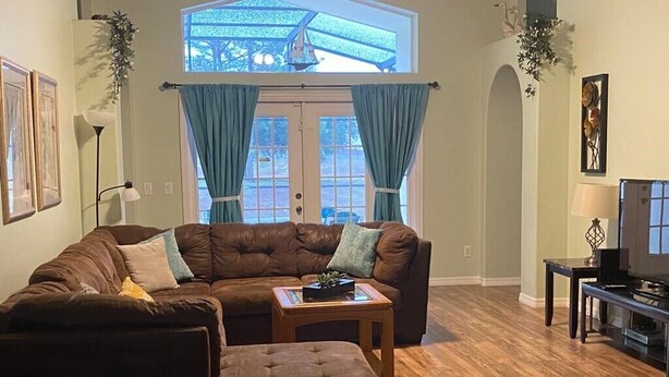 A living room with a brown leather sofa, a matching ottoman, a small wooden table with a plant, and a television on a stand. - Last-minute special, NEW beds/AC/pool heater/laminate floor, park view! (Kissimmee)