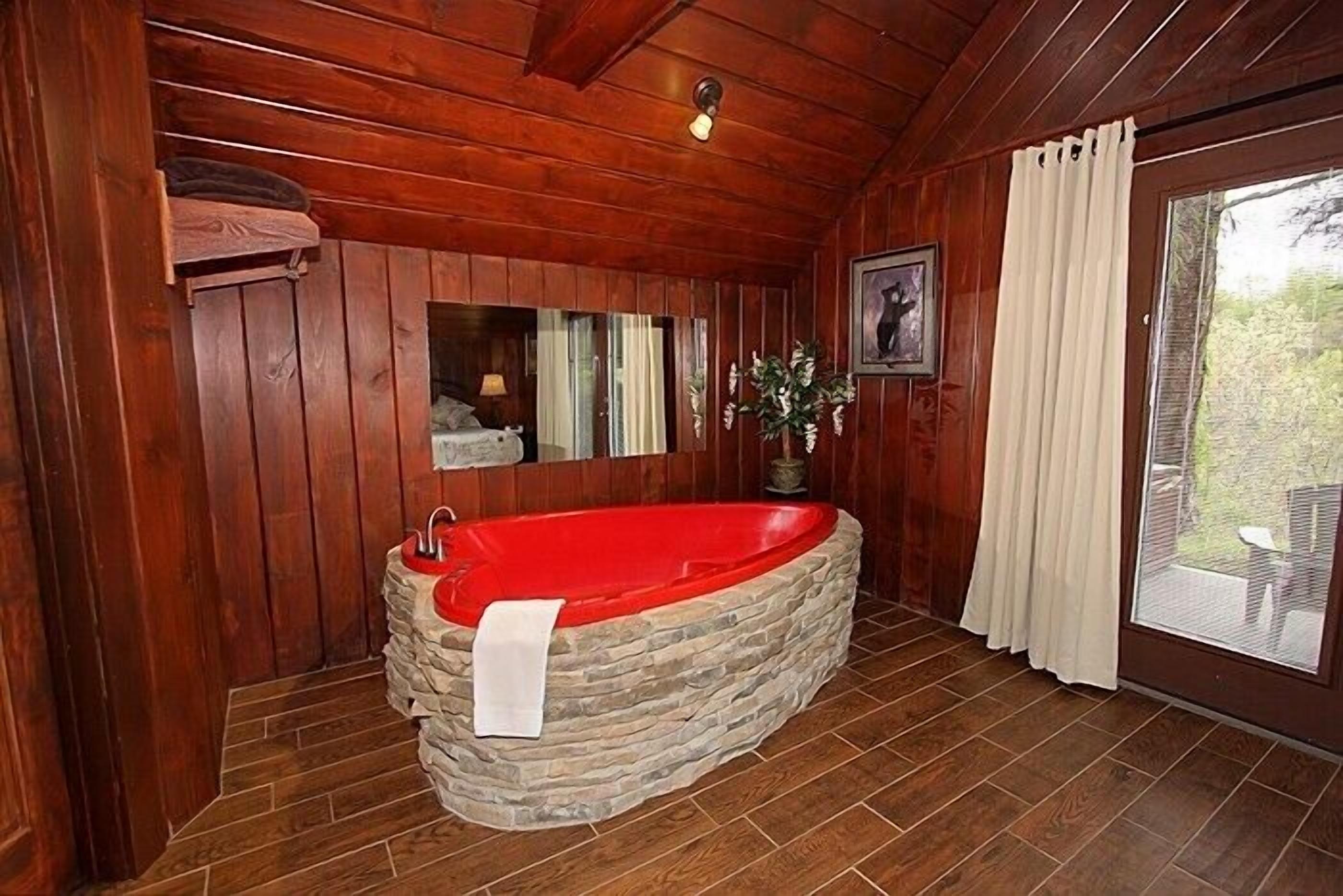 Une salle de bain rustique, avec des murs en bois, une baignoire en pierre, un miroir et une fenêtre agrémentée de rideaux.