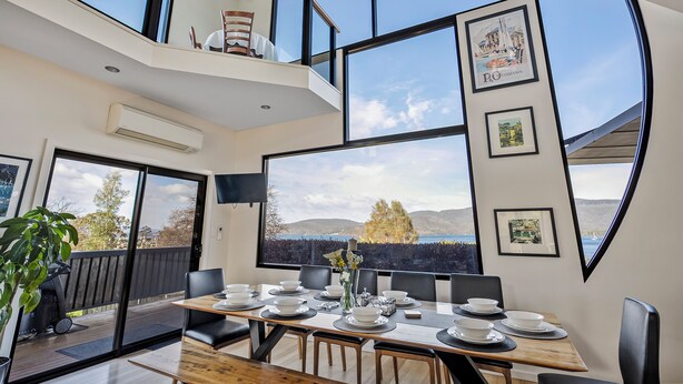 A dining area with a large window overlooking a scenic landscape, a wooden table set for dining, and framed pictures on the walls. - Quarry Hill Lookout ..... scenic views, luxurious amenities, relaxing atmosphere (Huon Valley)