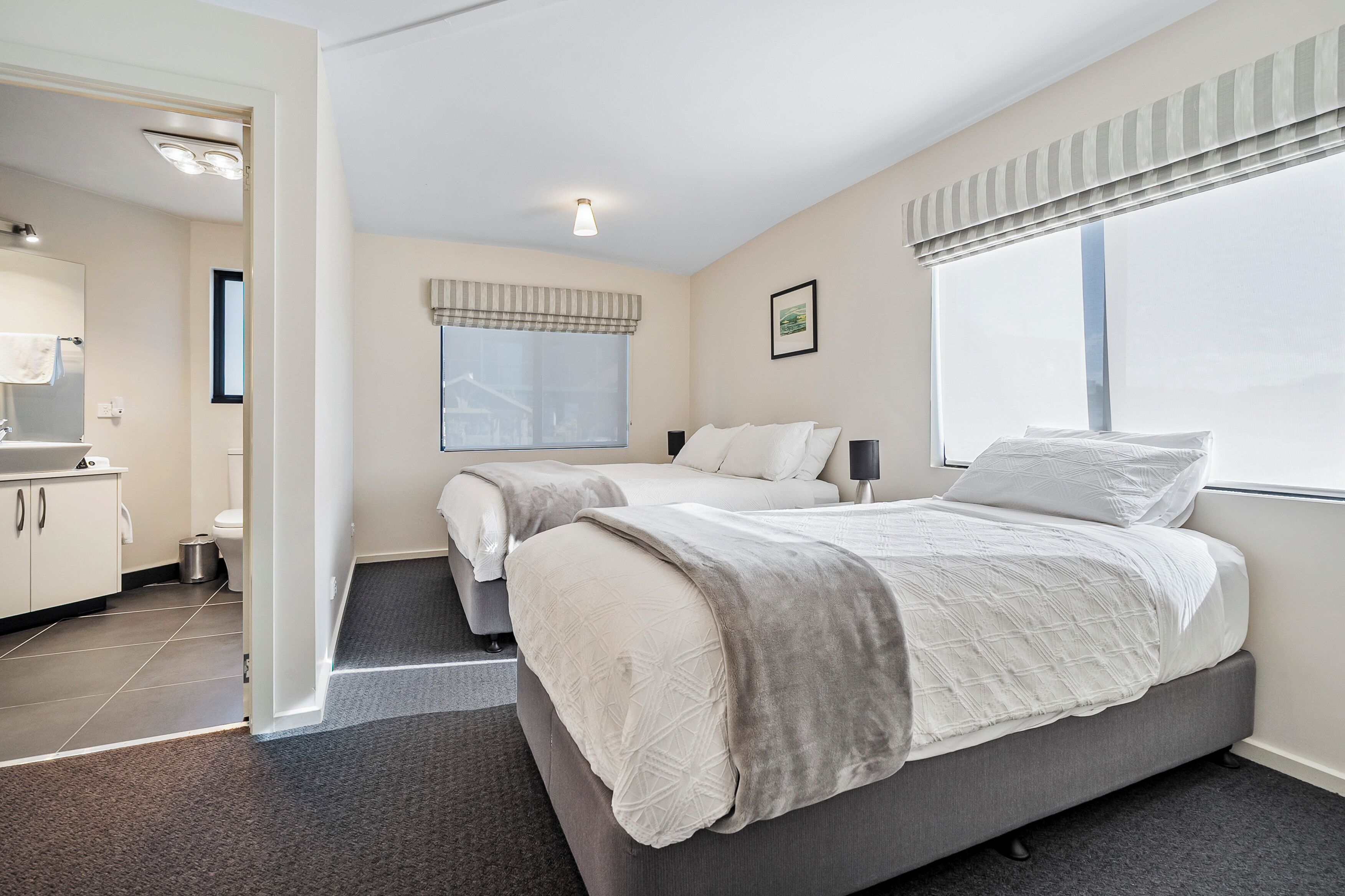 Una habitación de hotel moderna con una cama grande, dos mesitas de noche, un ventanal con persianas, un baño con inodoro y lavabo, y un pasillo que conduce a otra habitación.