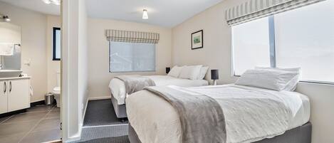 Una habitación de hotel moderna con una cama grande, dos mesitas de noche, un ventanal con persianas, un baño con inodoro y lavabo, y un pasillo que conduce a otra habitación.