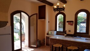 A kitchen with a white refrigerator, a microwave, and a stove. There are two stools and a wooden table. The kitchen has a tiled floor and a window with a view of greenery outside. - Villa with garden (Castel Volturno)