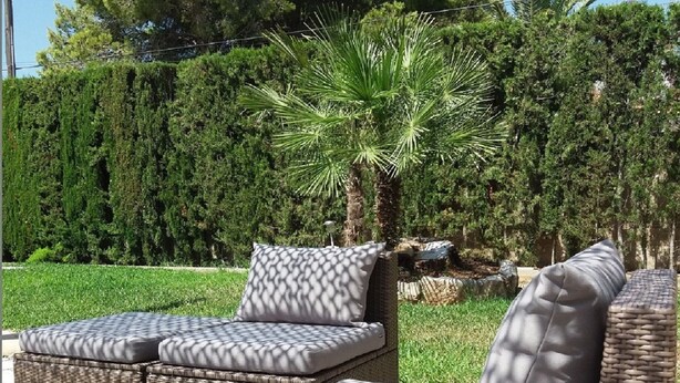Eine Sitzgruppe aus Rattan im Freien mit Kissen, umgeben von einem gepflegten Garten und Bäumen.