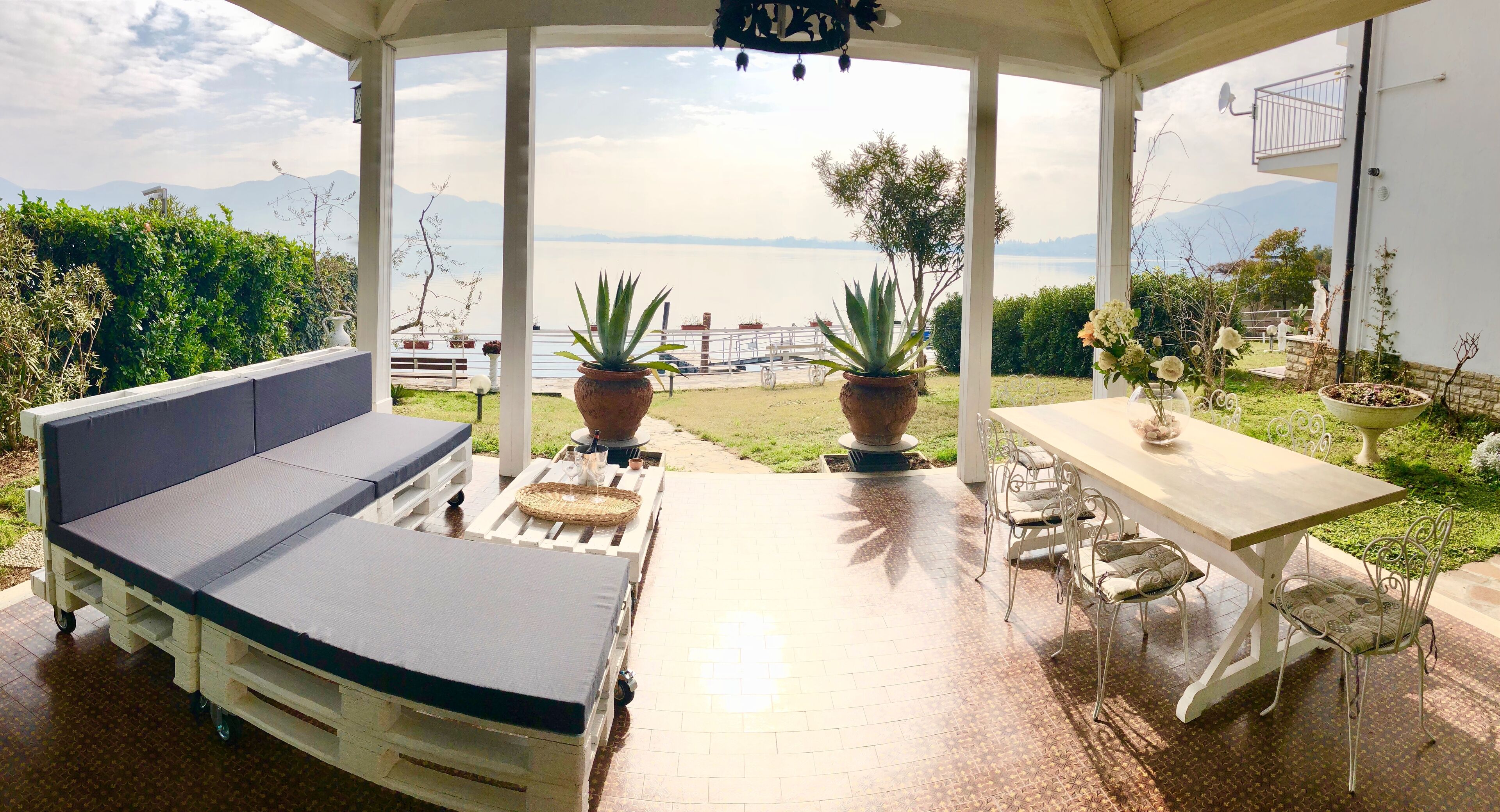 A covered outdoor seating area with a sofa and table, potted plants, and a view of the sea and mountains.