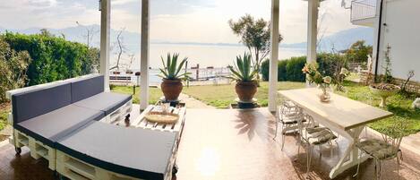 A covered outdoor seating area with a sofa and table, potted plants, and a view of the sea and mountains.