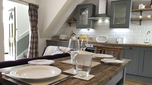 A dining area with a wooden table set for four, white plates, glasses, and a bottle of water. There is a chair, a purple sofa, and a kitchen in the background with grey cabinets and a white tiled backsplash. - The Cottage, a romantic getaway (Dunblane)