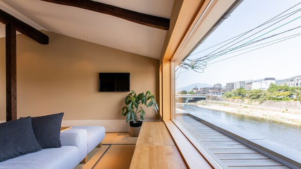 A modern interior with a sofa, a potted plant, and a view of a river through large windows. - Shirasagi Kyoto (Kyoto)
