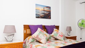 Een slaapkamer met een houten bed, twee nachtkastjes, een wandventilator en een schilderij van een zonsondergang boven de oceaan.
