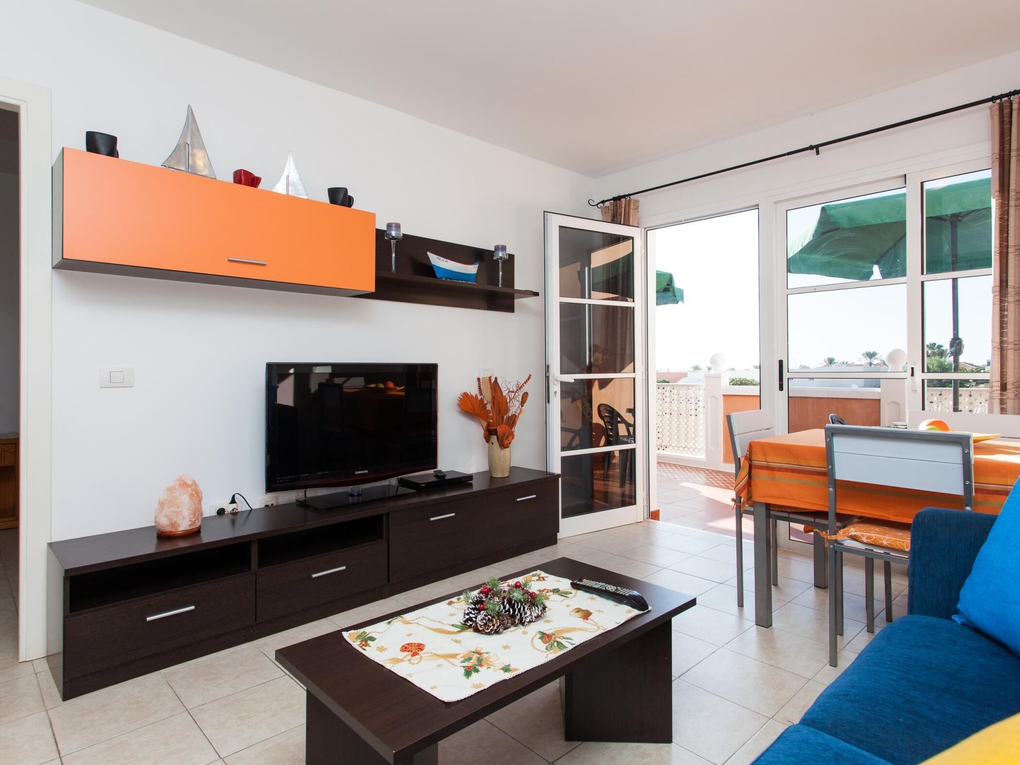 A modern living room with a flat-screen TV, dark wooden furniture, a dining area with a table and chairs, and a balcony with a view.