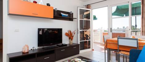 A modern living room with a flat-screen TV, dark wooden furniture, a dining area with a table and chairs, and a balcony with a view.