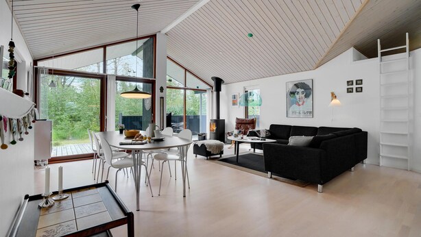 A modern living room with a large window, a black sofa, a dining table with chairs, and a fireplace. - Cozy home in ålbæk with WiFi (Ålbæk)