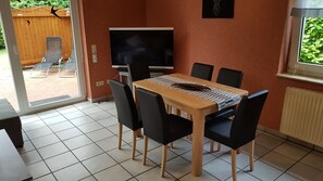Uma área de refeições com mesa, cadeiras, TV e janela com vista para área verde.