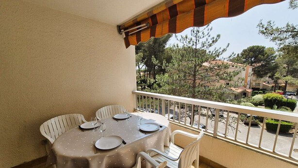 Un balcon avec une table dressée pour quatre personnes, des chaises blanches et une vue sur les arbres et les bâtiments.