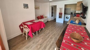 Une pièce de taille modeste comprenant un lit, une table à manger recouverte d’une nappe rouge, un ventilateur de plafond et un lave-linge.