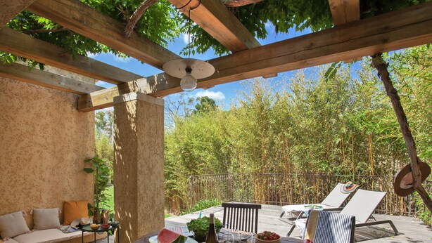 En träveranda med ett bord dukat för måltider, utomhusmöbler och en pergola ovanför.