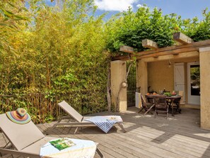 Una terraza de madera con dos sillones de relax, una mesa con mantel a rayas y una pérgola con una puerta.