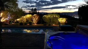 A swimming pool with a hot tub, surrounded by illuminated trees and bushes, with a sunset view in the background. - Perfect couple retreat paradise found at Casa De Luce (Kelowna)