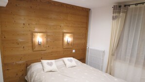 Ein Schlafzimmer mit einem hölzernen Kopfteil, zwei Wandleuchten, einem Bett mit weißer floraler Bettdecke und einem Fenster mit durchsichtigem Vorhang.