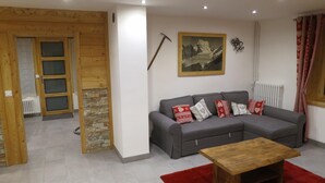 Ein Wohnzimmer mit einer grauen Eckcouch, einem Holztisch und einem roten Teppich. Eine Wand aus Stein, eine Holztür und eine Heizung sind ebenfalls vorhanden.