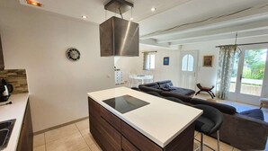 Eine moderne Küche mit Kochinsel, ein Essbereich mit einer Couch und ein Blick ins Freie durch ein großes Fenster.