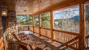 A wooden deck with a stone wall, wooden chairs, and a view of trees and mountains.