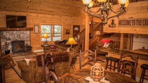 A cozy cabin interior with a stone fireplace, wooden furniture, a chandelier, and a welcome sign.