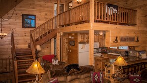 A cabin interior with a wooden staircase, a cozy living area with a sofa and lamps, a kitchen area, and a balcony with a cat.