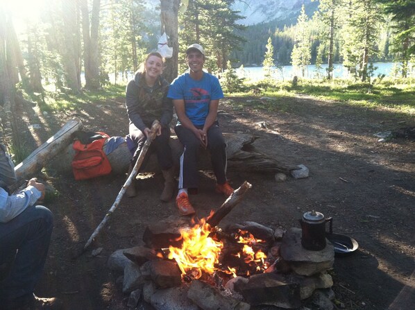 Two people sitting by a campfire in a forested area with a lake in the background.