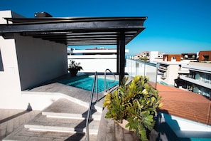 A rooftop pool area with a clear view of the sea, a covered seating area, and a staircase leading down.