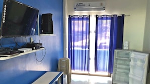 A room with a blue wall, a flat-screen TV mounted on the wall, a small table, a refrigerator, and a window with blue curtains.