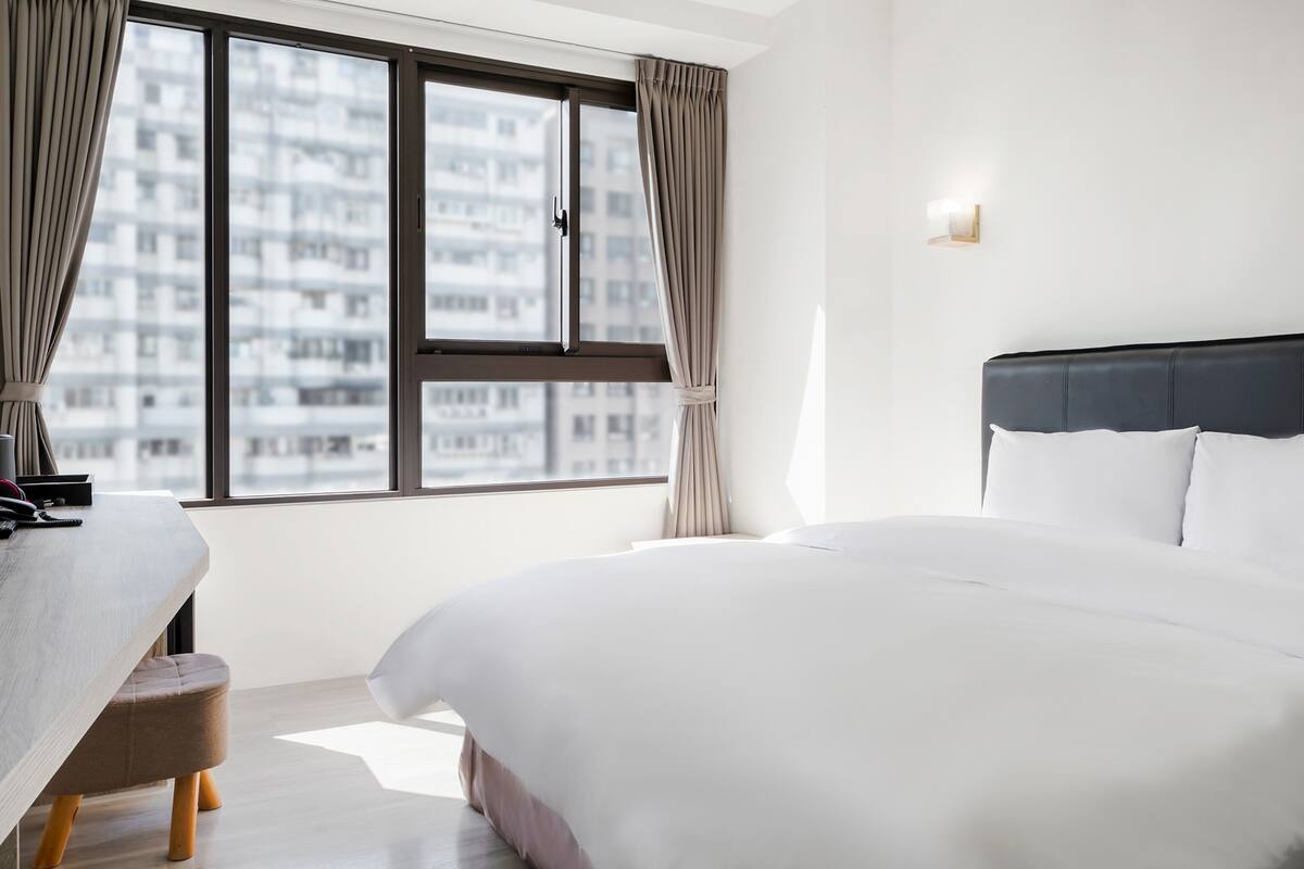 A modern hotel room with a large bed, a desk, and a view of a cityscape through a large window.