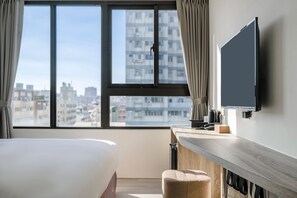 Habitación de hotel con cama, escritorio con televisor de pantalla plana, ventana con cortinas y vista a la ciudad.
