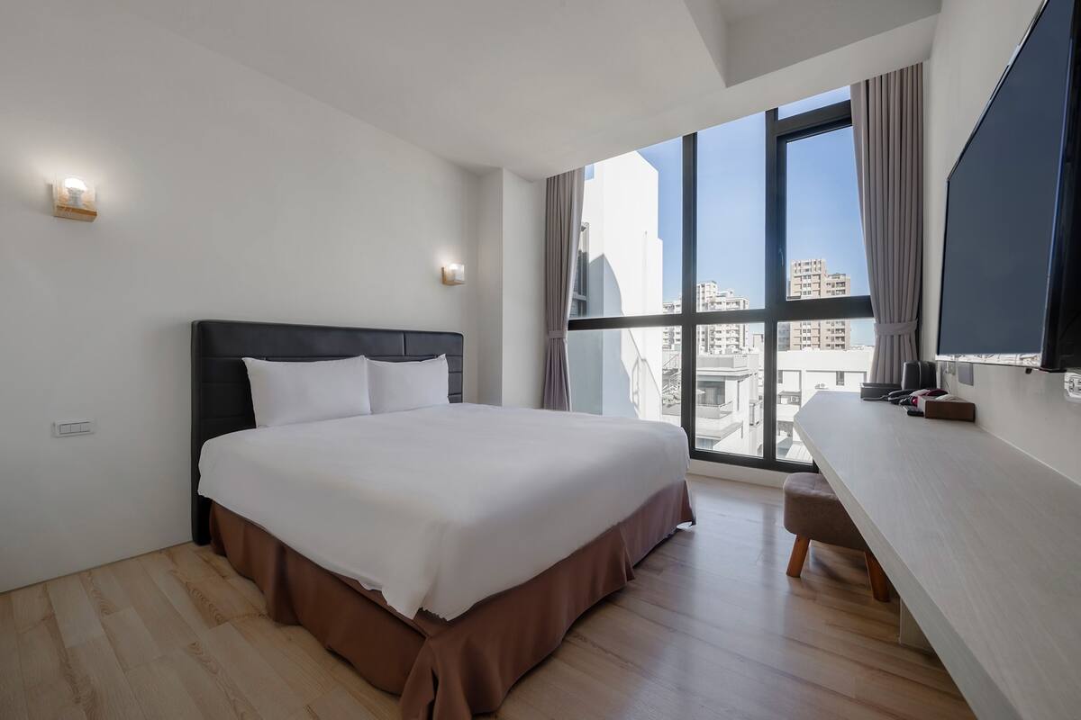 A modern hotel room with a large bed, a desk, and a large window offering a city view.